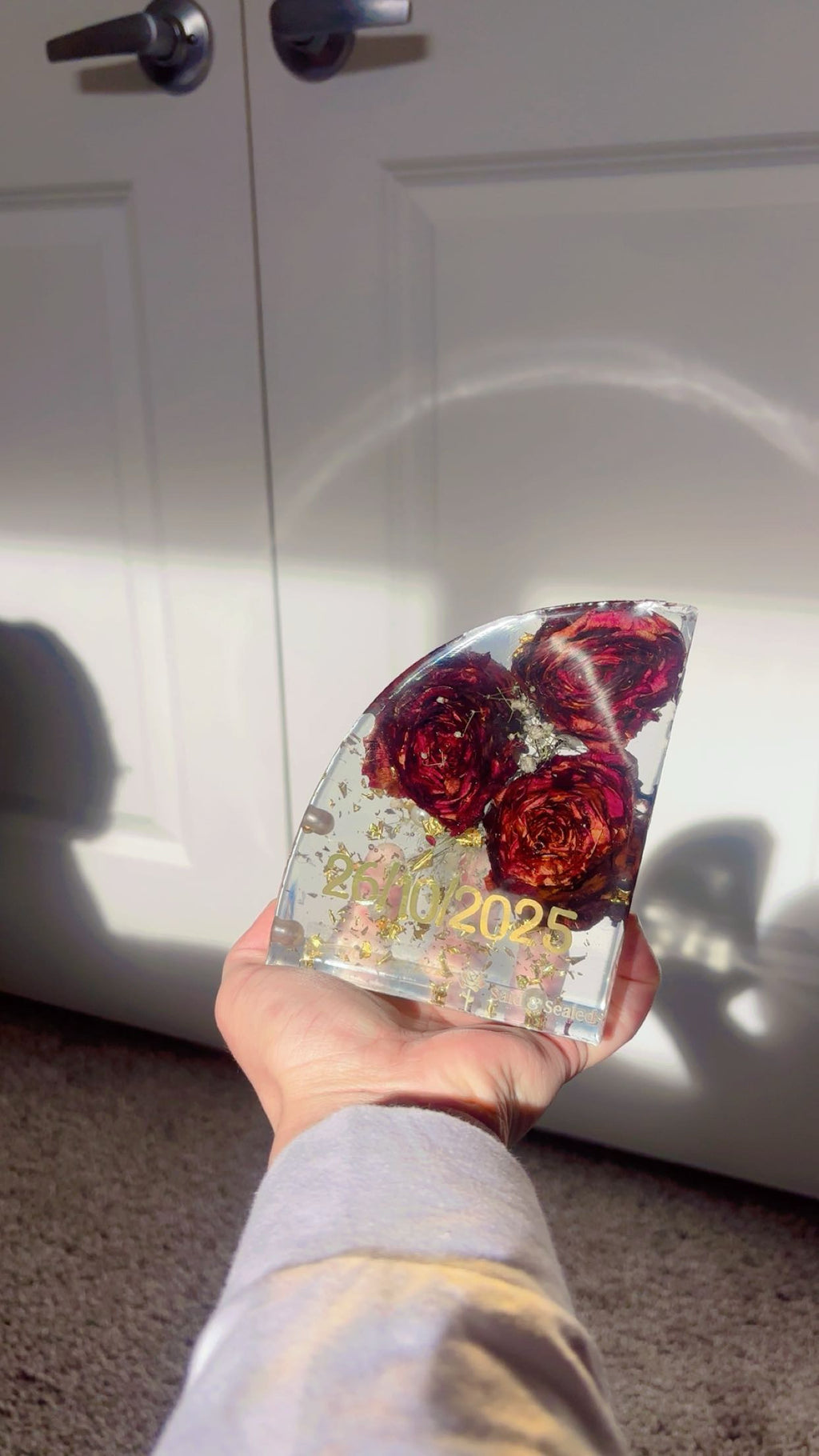 Hand holding a resin bookend with roses preserved inside and personalized with date 26,10,2025' text against a white door background